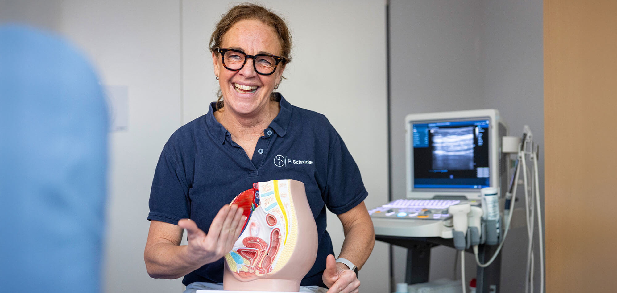Eine Physiotherapeutin im blauen Polo-Shirt erklärt mit einem anatomischen Beckenbodenmodell eine Untersuchungssituation und lächelt freundlich in Richtung Patientin.